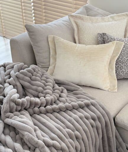 Paveikslėlio ALT žymė: Pilkas dvipusis dirbtinio kailio pledas ant sofos, jaukus ir šiltas interjero.